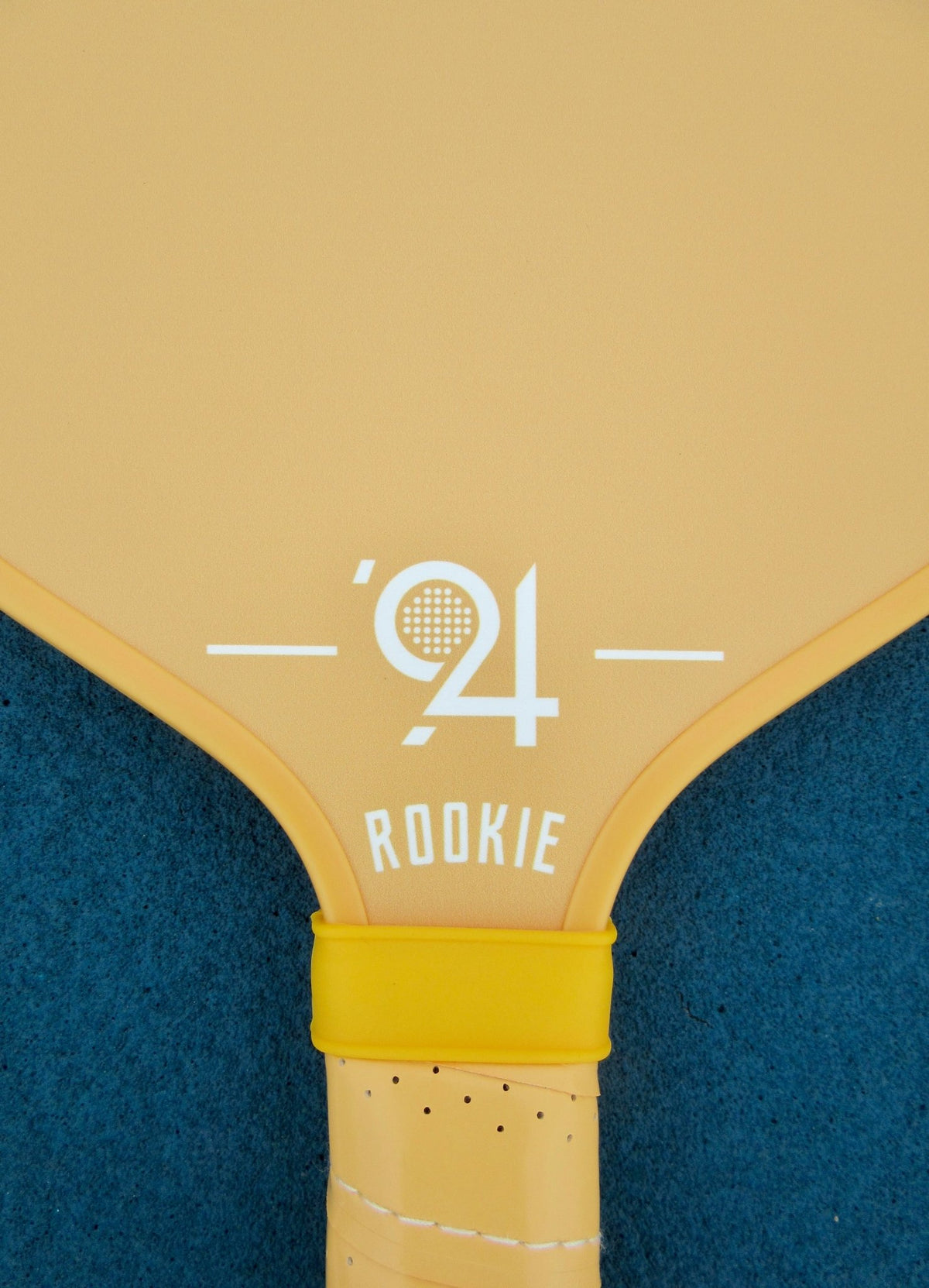 Rookie Youth Pickleball Paddle - Solara '94 - Pickleball Paddle - Close-up of a pickleball paddle featuring a pale yellow blade with white printed text reading "'94 ROOKIE" and a bright yellow wrist strap, set against a dark blue textured background.