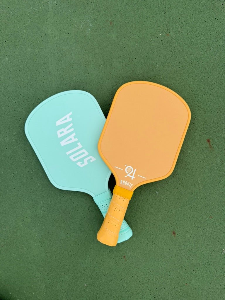 Rookie Youth Pickleball Paddle - Solara '94 - Pickleball Paddle - Two pickleball paddles, one mint green with white 'SOLARA' branding and one orange with a yellow handle, arranged on a green textured surface.