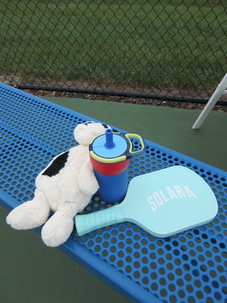 Rookie Youth Pickleball Paddle - Solara '94 - Pickleball Paddle - A light blue paddle-shaped item with the word 'SOLARA' printed on it, placed on a blue bench at an outdoor court with a stuffed animal and a blue, red, and yellow cup nearby.