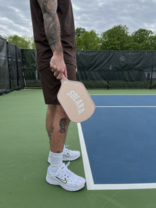 Tattooed player holding Solara '94 Gum pickleball paddle at the baseline of an outdoor court wearing white Nike sneakers
