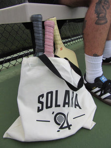 Solara '94 canvas tote bag courtside holding Legacy Collection paddles with visible Midnight, Gum, and Butter grips beside player wearing Adidas Sambas